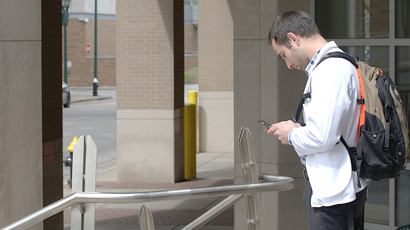 Man in a white lab coat with backpack looks down at phone.