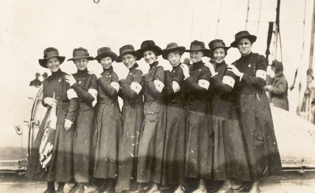 group of women standing one behind the other in a line in dark dresses and dark hats with nurse armbands and their hands are placed on the person in front of them to show off their arm bands