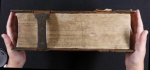 A pair of hands holds a large, closed antique book with metal clasps, viewed from the fore-edge against a black background.