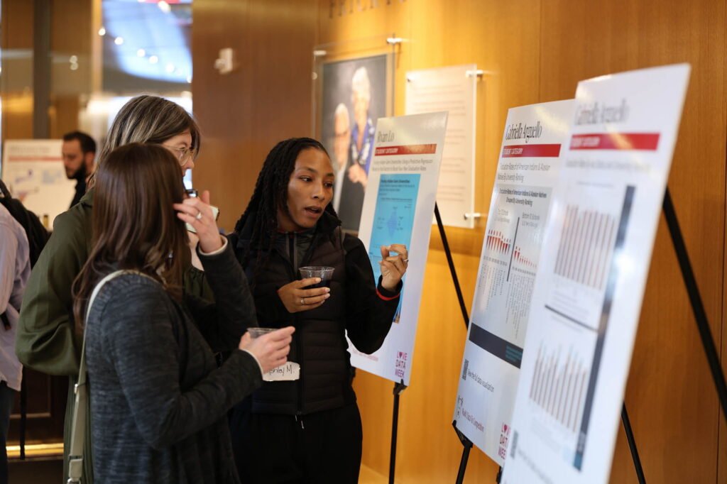 Three people stand in a hallway looking at research posters on display stands.