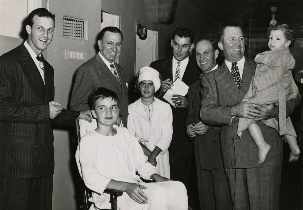 A group of men in suits surrounding a child sitting in a wheelchair and another child standing in a robe, with one man holding a toddler.