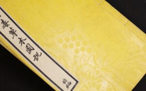 Yellow textured book cover with floral and grape patterns, featuring a white label with black Japanese calligraphy, partially visible against a black background.