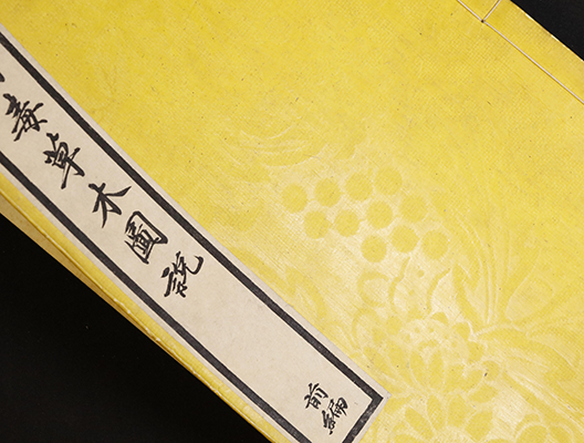 Yellow textured book cover with floral and grape patterns, featuring a white label with black Japanese calligraphy, partially visible against a black background.
