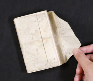 A hand lifts the cover of an old, stained book with a light-colored, possibly vellum or parchment binding, set against a black background.